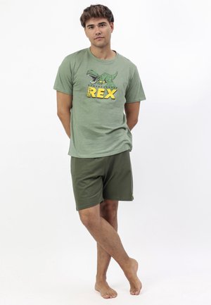 Joven de pie descalzo, vistiendo pantalones cortos verde oliva y una camiseta a juego con un dibujo animado de un dinosaurio y el texto "Nervous Rex".