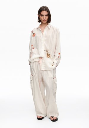 Mujer con cabello corto y oscuro, vestida con una camisa blanca suelta y pantalones con bordados florales y costuras rojas, de pie con las manos en los bolsillos.