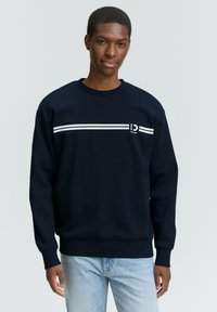 Sudadera azul marino hecha de un tejido suave, con una franja horizontal blanca y un logo en la parte frontal, y puños y cuello acanalados.