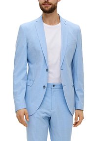 Man in een lichtblauwe blazer en broek met een wit T-shirt met ronde hals, staand tegen een witte achtergrond.