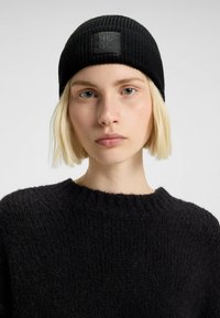 Cuffia nera lavorata a maglia con texture a coste e un patch quadrato con il logo "HUGO". Indossata con un maglione nero texturizzato.