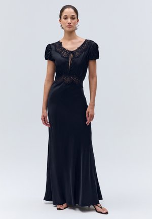 Femme portant une longue robe noire avec des détails en dentelle et des manches bouffantes, debout sur un fond uni gris clair.