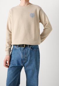 Persona che indossa una felpa beige con logo blu a forma di globo e jeans blu con cintura nera, in piedi di fronte a uno sfondo bianco.
