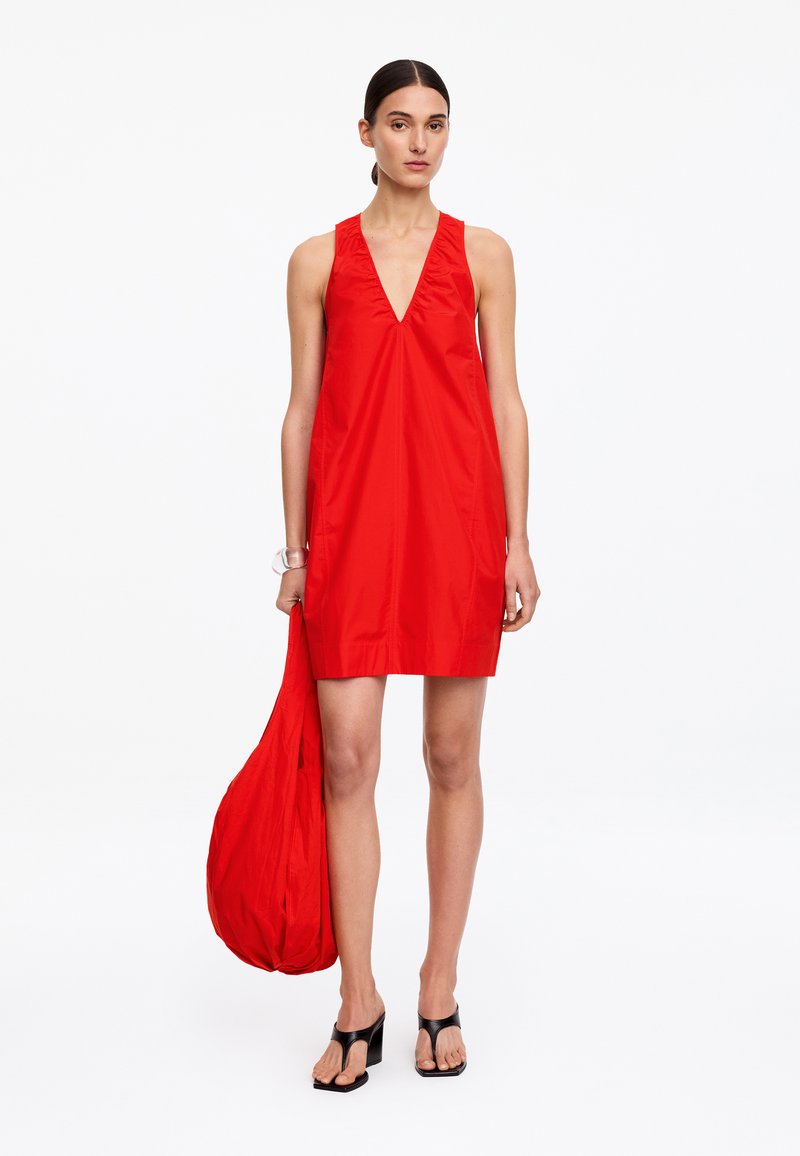 Femme portant une robe rouge sans manches à col en V, tenant un grand sac rouge assorti et des sandales à talons noires, debout sur fond blanc.