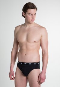 Zwarte briefs met een witte logo band, gemaakt van rekbare stof, met een aansluitend ontwerp en een gladde textuur, gedragen door een model.