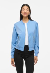 Sinine pommijakk, millel on lukk ees, soonikvarruks ja kaks lukuga küljetaskut. Sile kangas, lühike disain ja minima listlik stiil.