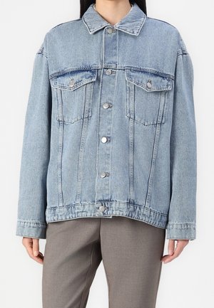 Lichtblauw spijkerjack met knoopsluiting en borstzakken, gedragen over grijze broek, getoond van nek tot heupen.