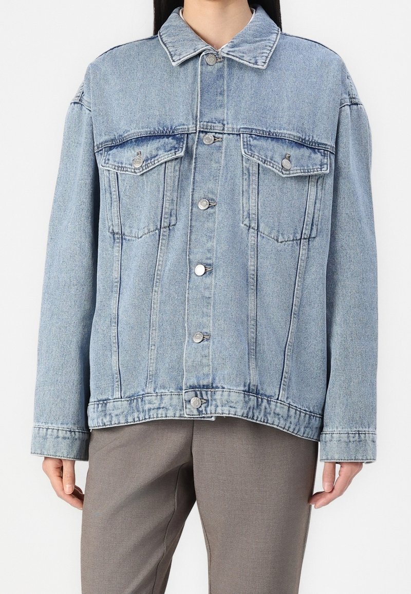 Veste en denim bleu clair avec fermeture à boutons et poches poitrine portée sur un pantalon gris, visible du cou aux hanches.