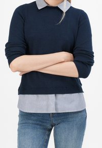 Pull en maille bleu marine avec poignets retroussés, porté sur une chemise à col rayée. Le bas a un ourlet droit, assorti à un jean bleu.