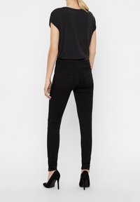 Jeans ajustados negros con una cintura de altura media y bolsillos traseros, combinados con una blusa negra suelta y tacones altos negros. Fondo liso.