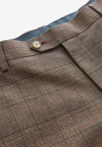 Gros plan d’un pantalon marron à carreaux avec une taille boutonnée et des passants de ceinture sur fond blanc.