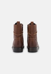 Bottines en cuir marron à texture lisse, avec une sangle décorative, une fermeture éclair arrière et un talon noir légèrement surélevé.