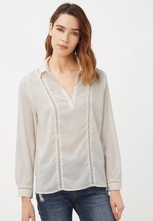 Mujer con cabello castaño hasta los hombros que lleva una blusa blanca con detalles de encaje vertical, mangas largas y jeans azules rotos.