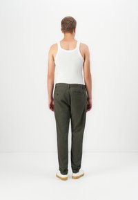 Homem em pé, de costas, vestindo uma t-shirt branca, calças verde-escuro e ténis brancos, contra um fundo branco liso.