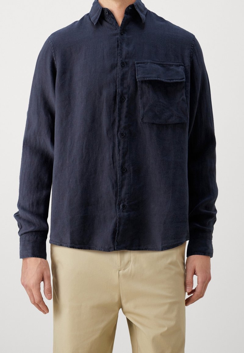 Homme portant une chemise bleu foncé à manches longues avec une poche poitrine et un pantalon beige sur un fond uni.
