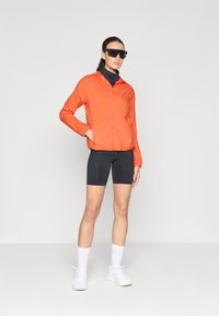 Orange lättviktsjacka med dragkedja fram, kombinerad med svarta shorts. Tillbehören inkluderar mörka solglasögon och vita sportskor med grå sulor.