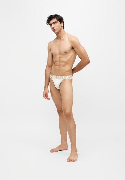 Jeune homme debout pieds nus portant un slip blanc avec une ceinture à motif doré sur un fond clair uni.