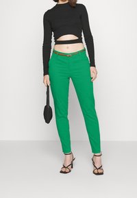 Kobieta w czarnej, długiej bluzce na krótki rękaw, zielonych, wysokich spodniach z brązowym paskiem, czarnych sandałach na obcasie, trzymająca czarną torebkę.