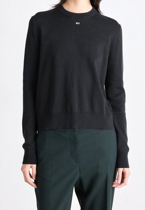 Person iført en sort, langærmet, rundhalset sweater med en lille logo på brystet og mørkegrønne skræddersyede bukser.