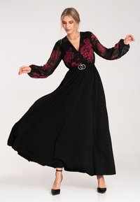 Sukienka maxi z czarną, zwiewną spódnicą, przezroczystymi długimi rękawami w burgundowy kwiatowy wzór oraz srebrnym paskiem z pierścieniem w talii.