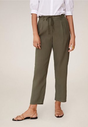 Kobieta w oliwkowo-zielonych spodniach ze sznurkiem, białej bluzce z bufkowymi rękawami oraz jasnobrązowych sandałach na płaskiej podeszwie, stojąca na jednolitym tle.