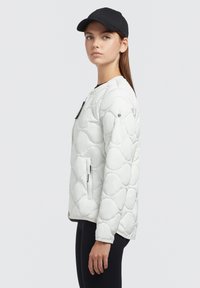 Hvid quilted jakke med rund krave, sidelommer med lynlås og en glat tekstur, suppleret af en sort kasket.