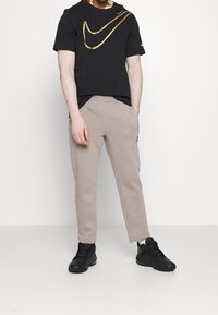 Muž v černém tričku Nike s velkým zlatým swooshem, béžových teplákových kalhotách a černých teniskách, stojící s rukama v kapsách.