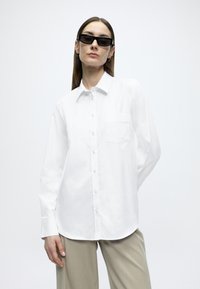 Λευκό πουκάμισο με κουμπιά, μυτερό γιακά, μακριά μανίκια και θήκη στο στήθος. Λείο ύφασμα με καθαρή σιλουέτα, συνδυασμένο με μπεζ παντελόνι.