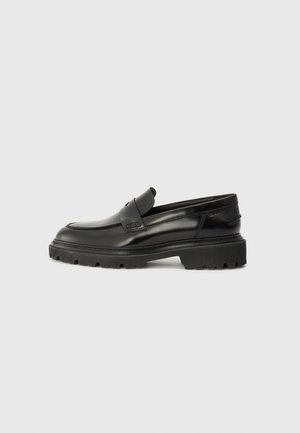 Scarpe slip-on in pelle nera con punta arrotondata, suola spessa e testurizzata, e dettagli di cucitura discreti. Presentano un piccolo logo sul retro.