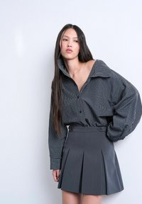 Femme aux longs cheveux noirs portant une chemise oversize à carreaux noirs et blancs et une jupe plissée grise taille haute sur un fond uni.