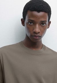 Hnědý bavlněný tričko s kulatým výstřihem, které má měkkou strukturu a minimalistický design. Model stojí čelem k fotoaparátu na neutrálním pozadí.