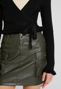 Zwarte geribbelde wikkeltrui met strikceintuur en gerimpelde zoom, gecombineerd met een donkergroene leren cargo-minirok met klepzakken.