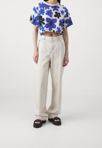 Camiseta blanca corta con patrones florales en azul y negro, combinada con pantalones sueltos color crema y sandalias de plataforma negras.