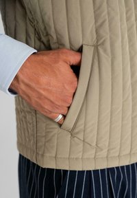 Hand with silver ring in pocket of beige quilted vest worn over light blue shirt and navy blue pinstriped pants.