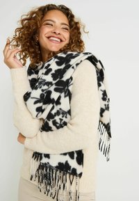 Bufanda con estampado floral en blanco y negro y bordes con flecos, hecha de tela suave y ligera, colocada sobre un suéter color crema.