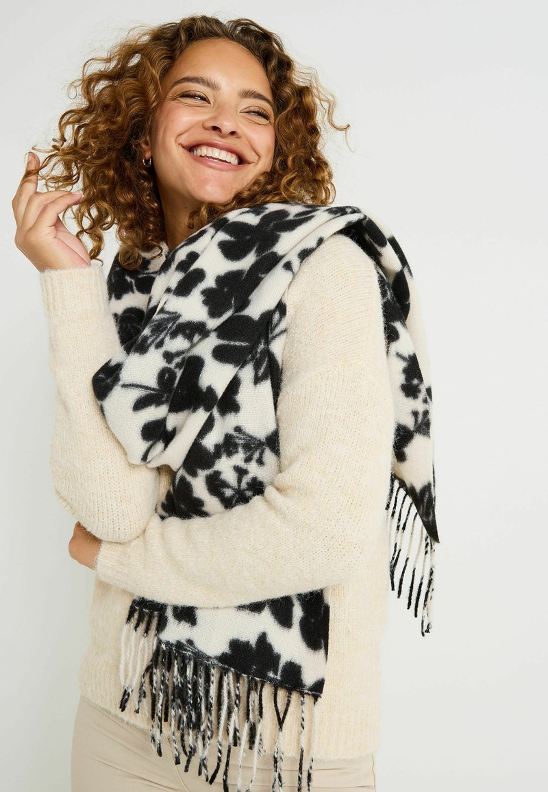 Bufanda con estampado floral en blanco y negro y bordes con flecos, hecha de tela suave y ligera, colocada sobre un suéter color crema.