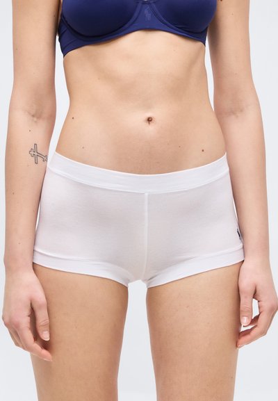 Γυναίκα που φοράει μπλε σουτιέν και λευκό εφαρμοστό σορτς, στέκεται με τα χέρια χαλαρά δίπλα στο σώμα της μπροστά σε λευκό φόντο.