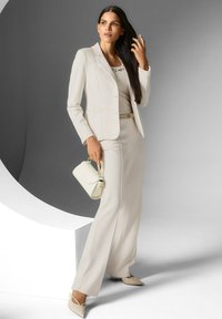 Vrouw in een crème kleurige blazer, bijpassende broek met brede pijpen en puntige hakken, die een kleine handtas vasthoudt en aan haar haar zit.