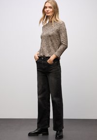 Beige Langarmshirt mit schwarzen Leopardenflecken, kombiniert mit schwarzen weit geschnittenen Jeans. Die Model trägt schwarze Stiefeletten. Einfacher Hintergrund.