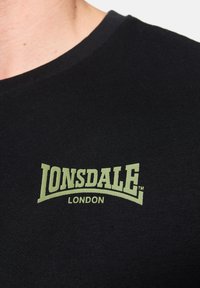 Fekete pamut póló bordás textúrával és kerek nyakkal. Jellemzője egy zöld logó, amelyen a "LONSDALE LONDON" felirat olvasható a mellkason.