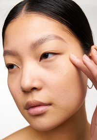 Gros plan sur un visage avec une peau lisse et claire ; un éclat subtil ; une boucle d'oreille en argent au nez ; et un doigt touchant la joue, indiquant l'application de soins de la peau.