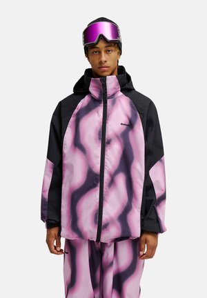 Junge Person trägt eine schwarz-rosa gemusterte Winterjacke und Hose mit lila Skibrille und schwarzer Mütze vor weißem Hintergrund.