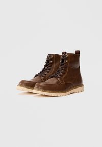 Braune Leder-Stiefeletten mit beigen Gummisohlen, runder Zehenform und Schnürung vorne. Mit Metallösen und einem Zugschlaufe hinten.