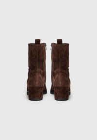 Botas de ante marrón hasta el tobillo con un tacón grueso, que presentan un cierre de cremallera y una tira en la parte posterior. Textura suave y diseño sencillo.