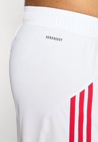 Shorts de sport blancs en tissu texturé, ornés d'un logo noir "AEROREADY" et de trois rayures verticales rouges sur le côté.
