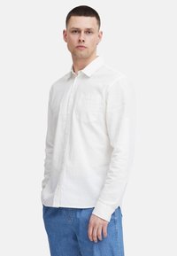 Weißes Langarmhemd aus leichtem Stoff mit Button-Down-Kragen, einer einzelnen Brusttasche und eher lockerer Passform, kombiniert mit blauen Jeans.