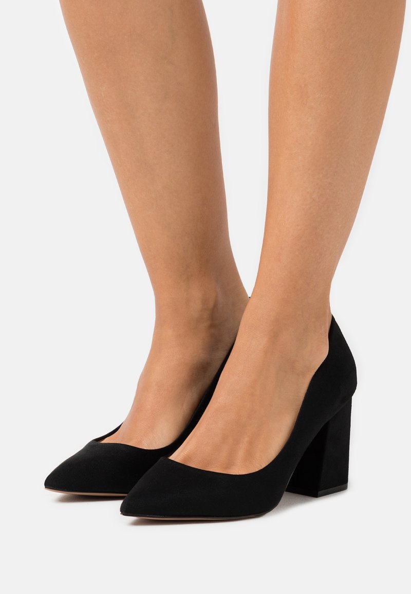 Schwarze Wildleder-Pumps mit hohen Absätzen, spitz zulaufenden Zehen und einem klobigen Blockabsatz. Glatte Textur und minimalistisches Design; keine sichtbaren Muster oder Akzente.