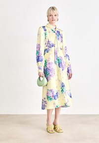 Florale midi-jurk in het geel, met paarse en groene hortensia-patronen, lange mouwen en een strikdetail bij de hals. Gekomplementeerd met een groene tas en gele hakken.