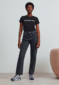 Černé tričko s nápisem "Calvin Klein Jeans", spárované s vysokými tmavými džíny a šedými teniskami. Jednoduchý, neformální outfit s čistými liniemi.