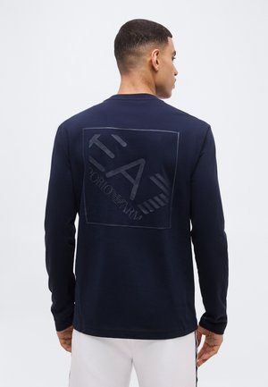 Άνδρας που φοράει μακρυμάνικο πουκάμισο σε ναυτικό μπλε με ανάγλυφο λογότυπο και κείμενο Emporio Armani στο πίσω μέρος, συνδυασμένο με λευκό παντελόνι.